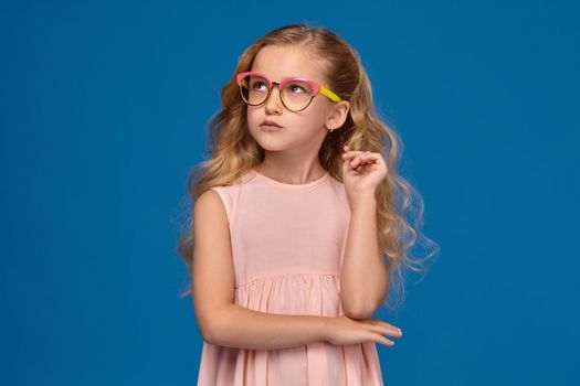 Pretty little girl in a pink dress and a fashionable glasses is looking up thoughtfully, standing on a blue background.