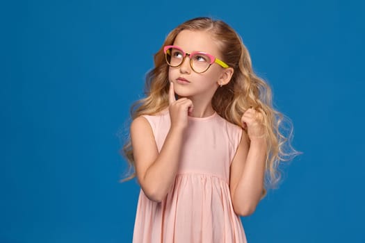 Beautiful little girl in a pink dress and a fashionable glasses is looking up thoughtfully, standing on a blue background.