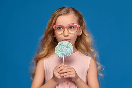 Pretty little girl in a pink dress and a fashionable glasses is licking a candy, standing on a blue background.