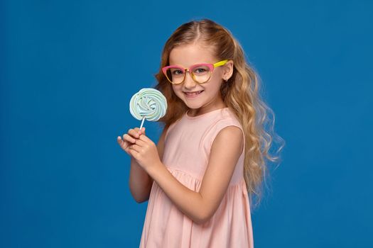 Nice little girl in a pink dress and a fashionable glasses is holding a candy and smiling, on a blue background.