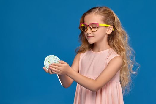 Charming little girl in a pink dress and a fashionable glasses is holding a candy, standing on a blue background.