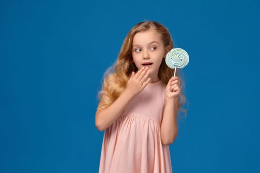 Nice little girl in a pink dress is holding a candy and covering her mouth with her hand, on a blue background.