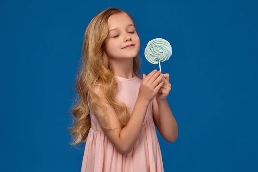 Beautiful little girl in a pink dress is holding a candy and looking on it, on a blue background.