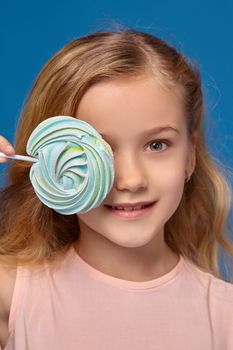 Gorgeous little girl in a pink dress closes her eye with a candy, standing on a blue background.