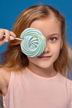 Pretty little girl in a pink dress closes her eye with a candy, standing on a blue background.