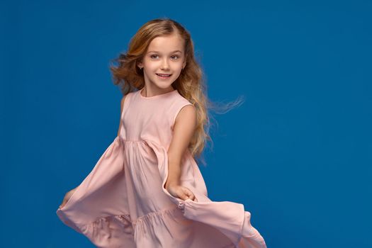 Pretty little girl in a pink dress, with a curly hair is dancing and posing, on a blue background.