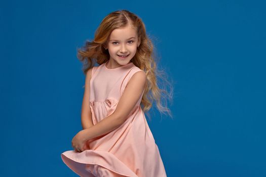 Pretty little girl in a pink dress, with a curly hair is spinning around, on a blue background.