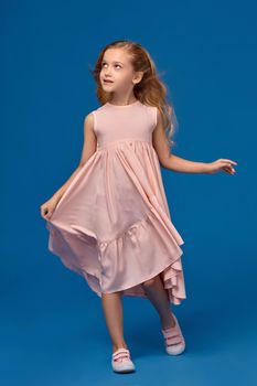 Nice little girl in a pink dress, with a curly hair looks away, on a blue background.