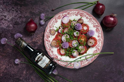 As,Belgium, May 10, 2022: tomato salad with purple onion rings dressed with Ponti balsamic dressing and olive oil with feta cheese and olives on a dark purple background, step by step recipe,flat lay