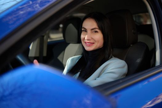 Attractive middle-aged dark-haired European successful woman, in formal wear, driving car and smiling a beautiful toothy smile looking at camera. People, Lifestyle, Urban life and Business concept