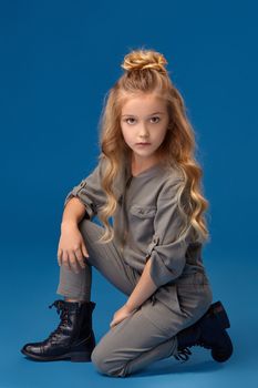 Little girl with a curly blond hair in a fashionable gray overall and black boots, set down on her knee, on a blue background