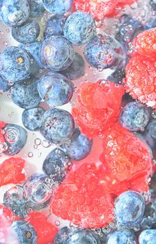 Blueberries and raspberries in liquid with bubbles. Colourful ripe bilberries and raspberries in water. Close-up of fresh berries in water background. Top view, flat design. Vertical macro image.