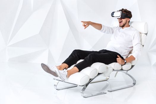 Excited young man is getting experience using VR-headset glasses of virtual reality gesticulating with his hands. A young man sits on a comfortable armchair in a room with white walls