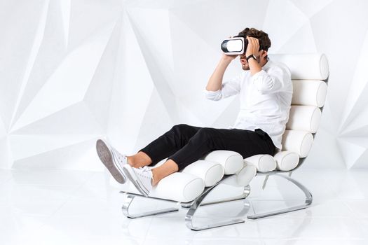 Excited young man is getting experience using VR-headset glasses of virtual reality gesticulating with his hands. A young man sits on a comfortable armchair in a room with white walls