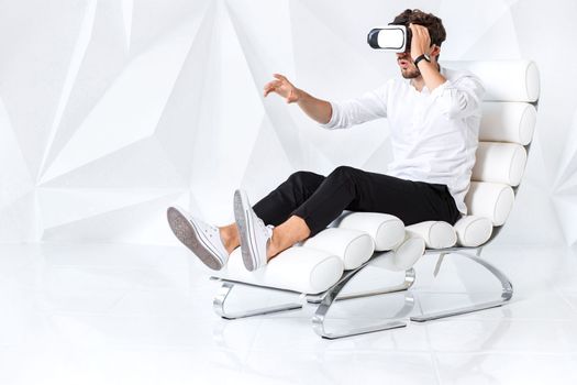 Excited young man is getting experience using VR-headset glasses of virtual reality gesticulating with his hands. A young man sits on a comfortable armchair in a room with white walls