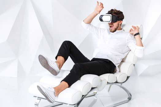 Excited young man is getting experience using VR-headset glasses of virtual reality gesticulating with his hands. A young man sits on a comfortable armchair in a room with white walls