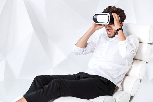 Excited young man is getting experience using VR-headset glasses of virtual reality gesticulating with his hands. A young man sits on a comfortable armchair in a room with white walls