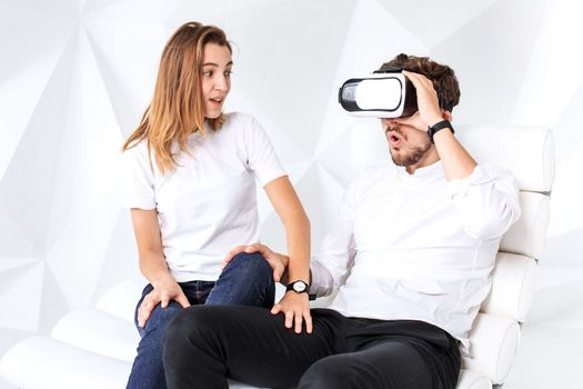 Couple having fun playing with virtual reality. A young man sits on a comfortable armchair in a room with white walls