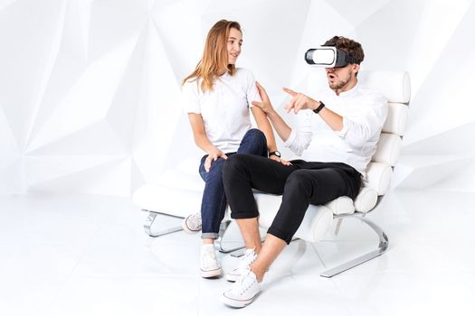 Couple having fun playing with virtual reality. A young man sits on a comfortable armchair in a room with white walls