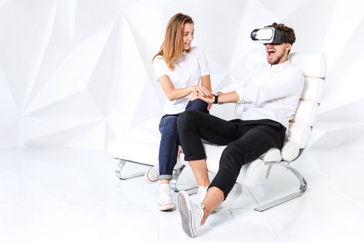 Couple having fun playing with virtual reality. A young man sits on a comfortable armchair in a room with white walls