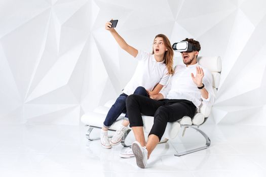 Couple having fun playing with virtual reality. A young man sits on a comfortable armchair in a room with white walls
