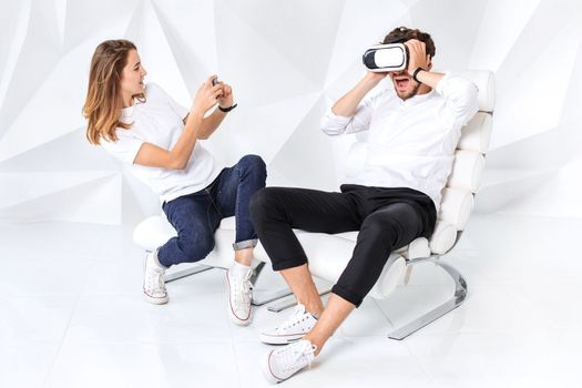 Couple having fun playing with virtual reality. A young man sits on a comfortable armchair in a room with white walls