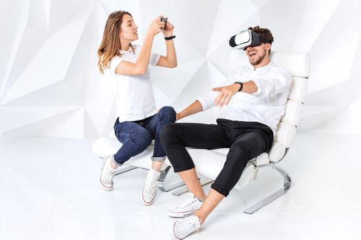 Couple having fun playing with virtual reality. A young man sits on a comfortable armchair in a room with white walls