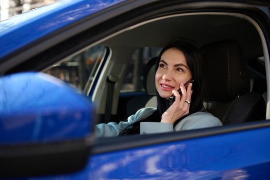 Close-up attractive stylish woman sitting in car on driver's seat, dressed in coat winter style, using mobile phone, talking with clients, plans business meeting. Successful businesswoman driving car