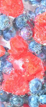 Blueberries and raspberries in liquid with bubbles. Colourful ripe bilberries and raspberries in water. Close-up of fresh berries in water background. Top view, flat design. Vertical macro image.