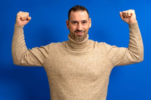 Latino man, happy, unshaven, strong, sporty, fitness student, 40s man in turtleneck showing biceps muscles on hand isolated on blue color background studio portrait