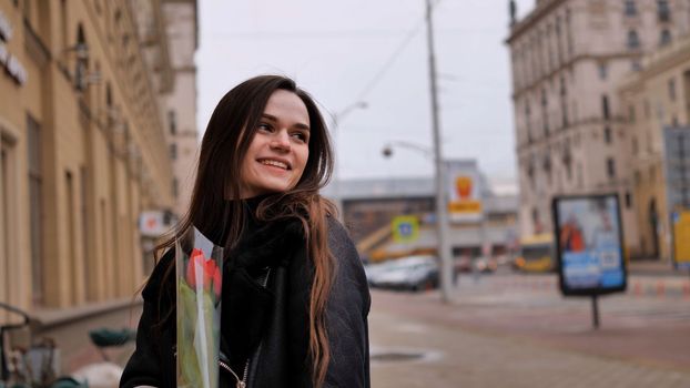 A happy brunette girl in a black jacket walks through the city in early spring