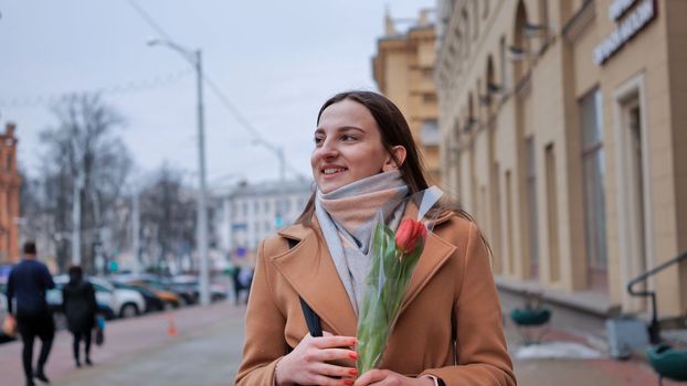 A happy brunette girl in a black jacket walks through the city in early spring