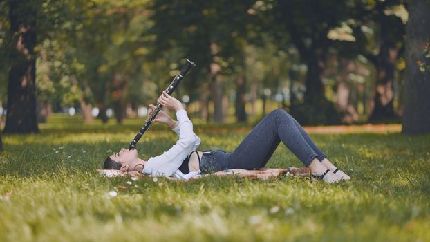 A girl plays the clarinet while lying on the grass in the park