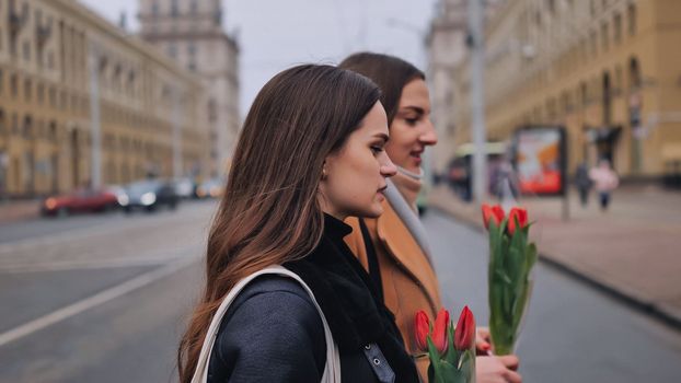 Two joyful friends in a jacket and coat are walking around the city with tulips