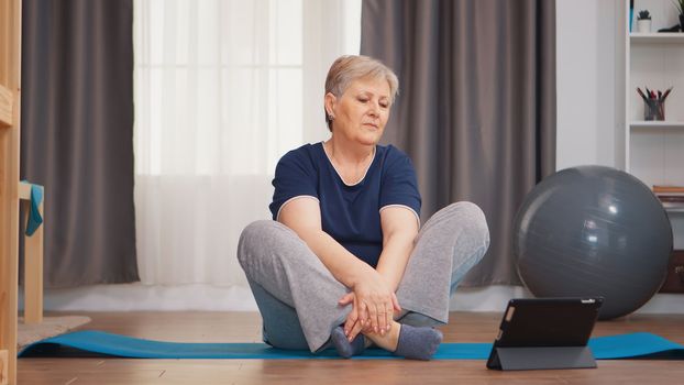 Senior woman warming up shoulder watching online tutorial sitting on yoga mat. Online learning and study, Active healthy lifestyle sporty old person training workout home wellness and indoor exercising
