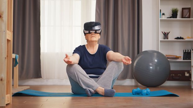 Senior woman meditating on yoga mat wearing vr headset. Active healthy lifestyle sporty old person training workout home wellness and indoor exercising