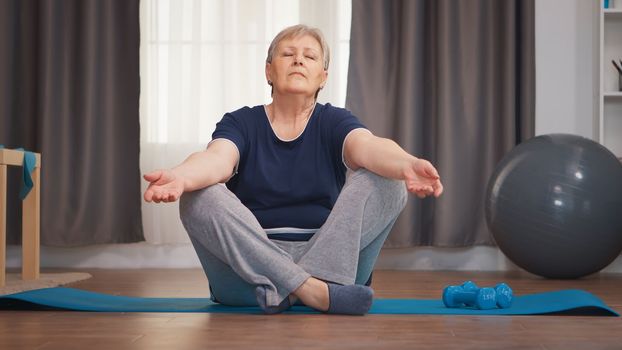 Senior woman meditating sitting on yoga mat in living room. Active healthy lifestyle sporty old person training workout home wellness and indoor exercising