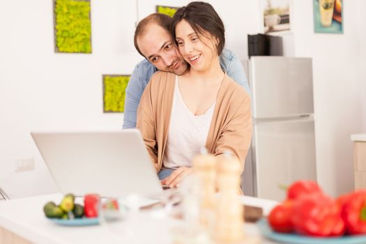 Smiling couple using laptop in kitchen with healthy vegetables on the table. Happy loving cheerful romantic in love couple at home using modern wifi wireless internet technology