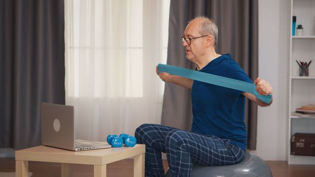 Senior man with vitality doing fitness exercise with resistance band watching online program. Old person pensioner healthy training healthcare sport at home, exercising fitness activity at elderly age