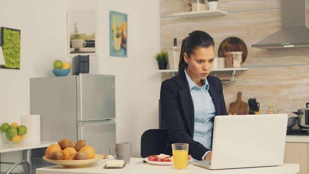 Business woman eating toasted bread with butter while working on laptop during breakfast. Concentrated business woman in the morning multitasking in the kitchen before going to the office, stressful way of life, career and goals to meet