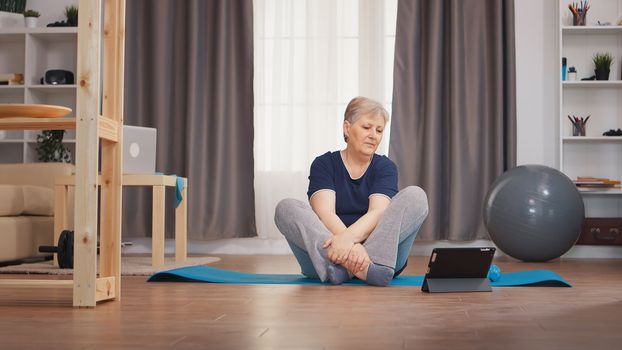 Senior woman stretching body watching online lesson sitting on yoga mat. Online learning and study, active healthy lifestyle sporty old person training workout home wellness and indoor exercising