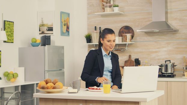 Young business woman in the kitchen having a healthy meal while talking on a video call with her colleagues from the office, using modern technology and working around the clock