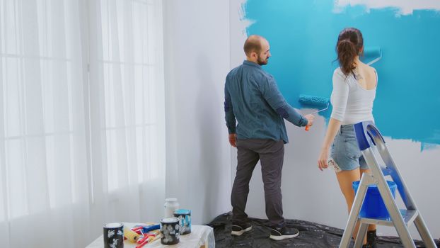 Caucasian couple painting wall with roller brush and blue paint. Apartment redecoration and home construction while renovating and improving. Repair and decorating.