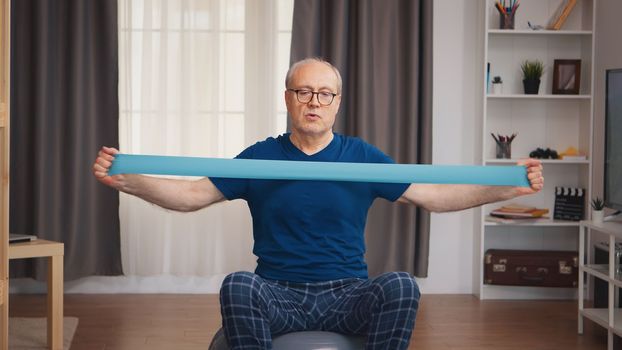Senior man on balance ball training with resistance band. Old person pensioner healthy training healthcare sport at home, exercising fitness activity at elderly age