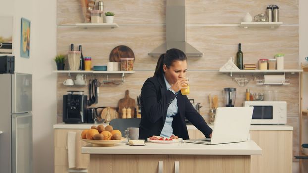 Business woman eating breakfast and working on laptop. Concentrated business woman in the morning multitasking in the kitchen before going to the office, stressful way of life, career and goals to meet