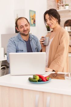Cute couple using laptop together in kitchen. Freelancer wife holding cup of coffee. Happy loving cheerful romantic in love couple at home using modern wifi wireless internet technology