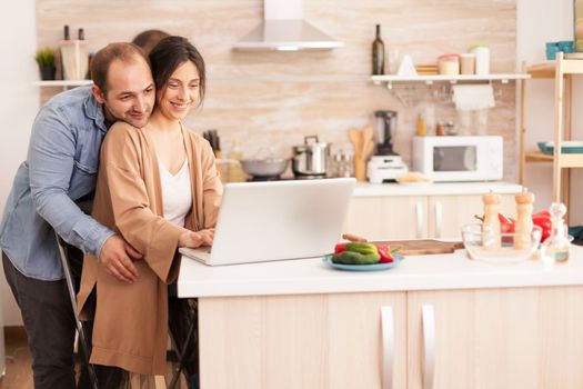 Cheerful couple using laptop in kitchen reading online recipe for breakfast. Happy loving cheerful romantic in love couple at home using modern wifi wireless internet technology