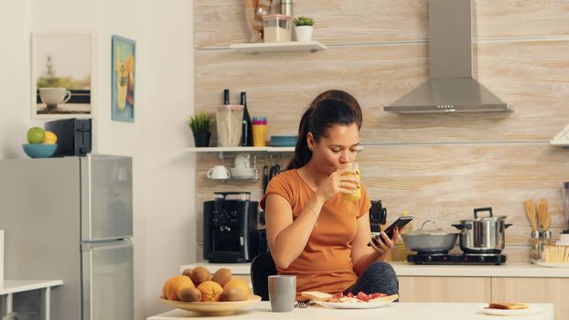 Woman browsing on phone at breakfast and drinking fresh orange juice with a healthy organic meal and a cup of coffee, using modern technology alone in the kitchen before going to work.