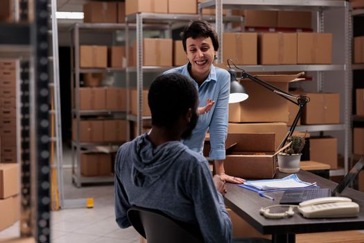 Storehouse workers having fun at job while preparing customers packages putting order in carton box, working in warehouse delivery department. Diverse team checking cargo stock on laptop computer