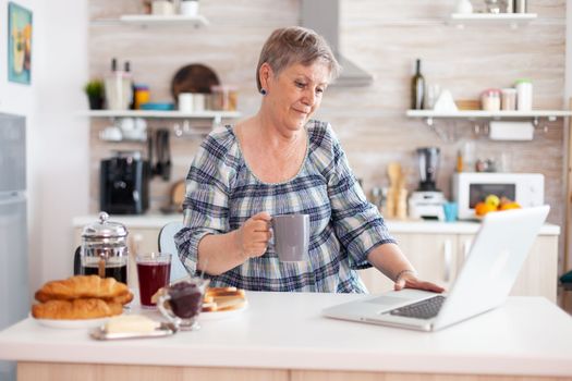 Senior businesswoman drinking coffee and working on laptop in kitchen during breakfast. Elderly retired person working from home, telecommuting using remote internet job online communication on modern